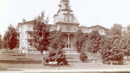 The Athenaeum Hotel built in 1881. In the foreground are very young trees, older wooden benches and an unpaved walk.