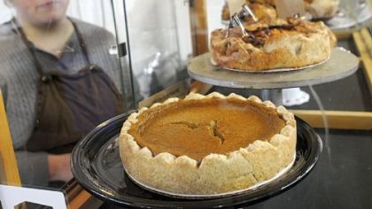 Sara Kelby Lewis, the owner/baker of Sustenance Rustic Craft Bakery, is seen behind the large glass case at her stand in the Pittsburgh Public Market.  The pie in the foreground is pumpkin.